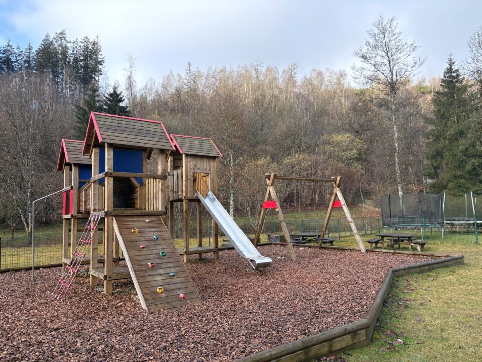 Jouer à l'intérieur comme à l'extérieur Une plaine de jeu avec toboggan, balançoires, et copeaux de bois au sol, le tout entouré de nature et d'arbres.