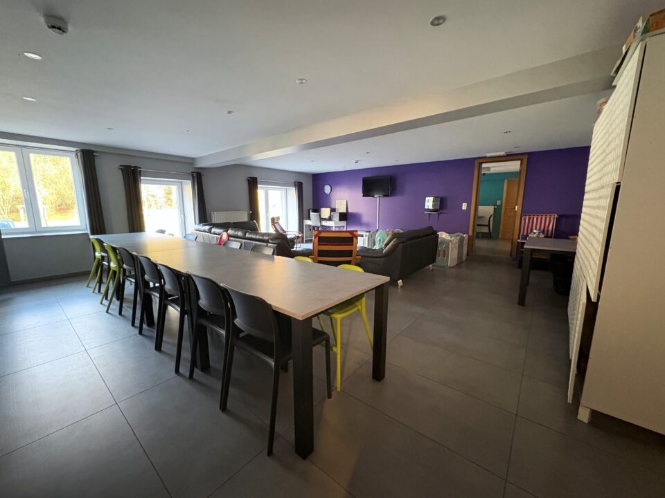 Tous à table ! Une photo d'une salle illuminée par de nombreuses fenêtres, au centre de la pièce se trouve une table avec de nombreuses chaises. En fond, on peut appercevoir un canapé.