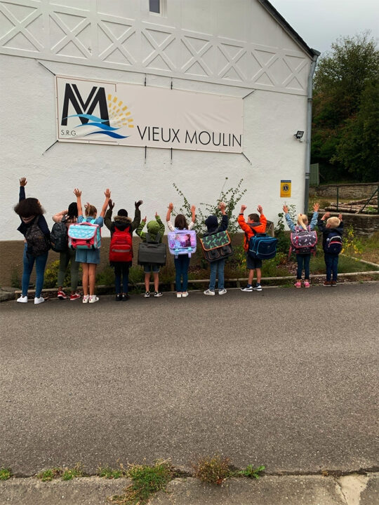 Un groupe de 10 enfants levant les bras, vus de dos, face à un mur blanc sur lequel figure le logo du Vieux Moulin