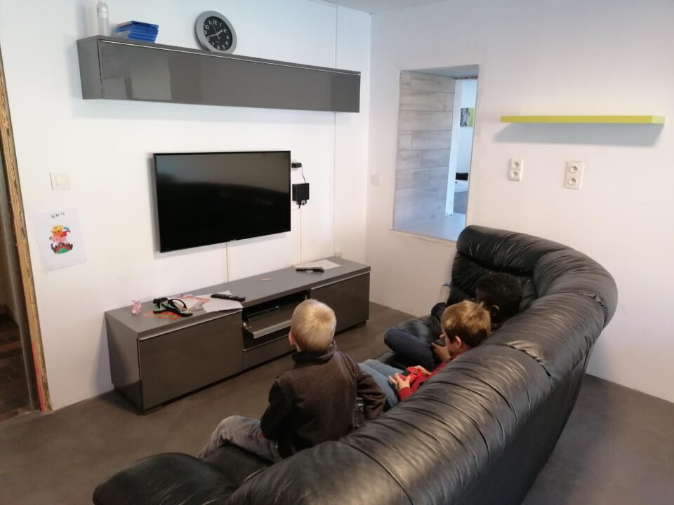 Une photo de trois enfants assis dans un canapé, tenant les manettes d'une console en main, faisant face à un écran plat accroché au mur.