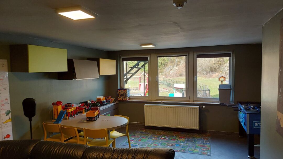 Une photo de l’intérieur du Vieux Moulin, montrant une salle avec un tapis de jeu au sol, des tables couvertes de jouets pour enfants, et des fenêtres vers l'extérieur montrant la cour intérieure bordée de verdure