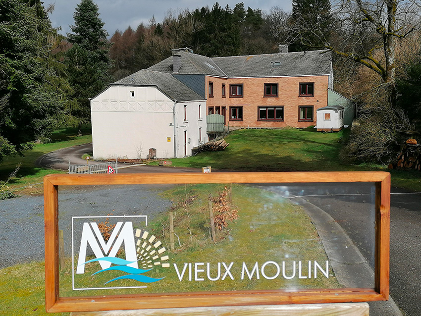 Une photo prise de loin du Vieux Moulin, avec en premier plan un panneau sur lequel est placé le logo du Vieux Moulin.